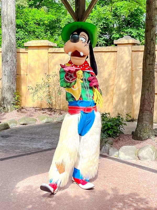 Goofy dressed in a fun cowboy outfit at Disneyland Paris in April, posing playfully in Frontierland, surrounded by trees and rustic scenery.