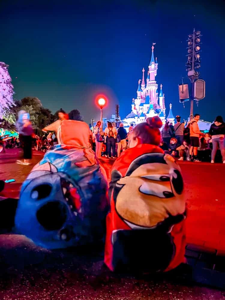 Disneyland Paris guests wrapped in character blankets sitting on the curb before the fireworks — a smart comfort tip for enjoying the best Disneyland Paris fireworks spot.