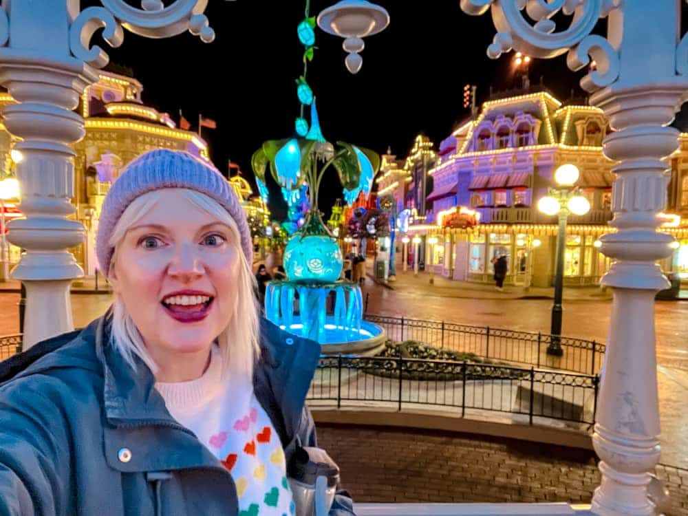 Disney blogger Carrie-Anne smiling on Main Street at Disneyland Paris during the evening with colourful lights behind her — planning ahead to enjoy the best Disneyland Paris fireworks spot.