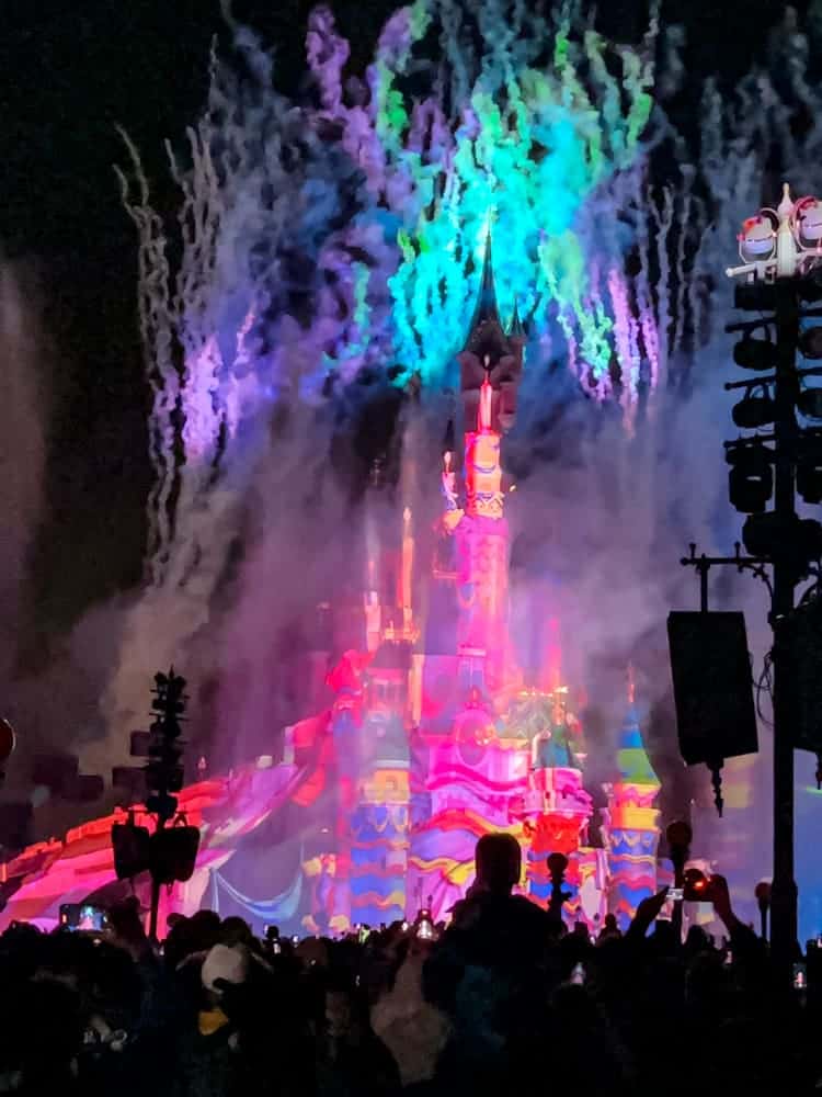 Fireworks bursting behind the castle with colourful projections during Tales of Magic at Disneyland Paris — showing a crowd watching from one of the best Disneyland Paris fireworks spots.