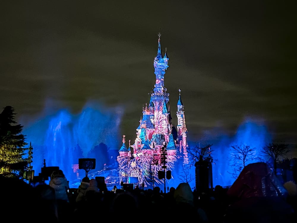 Castle lit with pink and purple forest projections at Disneyland Paris with silhouettes of guests watching the fireworks — an ideal example of the best Disneyland Paris fireworks spot on the Discoveryland entrance side for views and atmosphere.