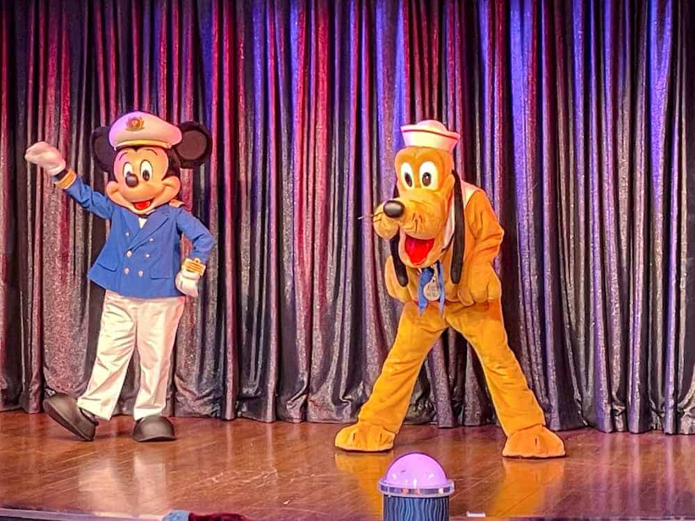 Mickey Mouse in a blue Disney Cruise Line captain’s uniform and Pluto in a sailor hat perform together on stage in front of a shimmering curtain during a Disney Cruise Line show.