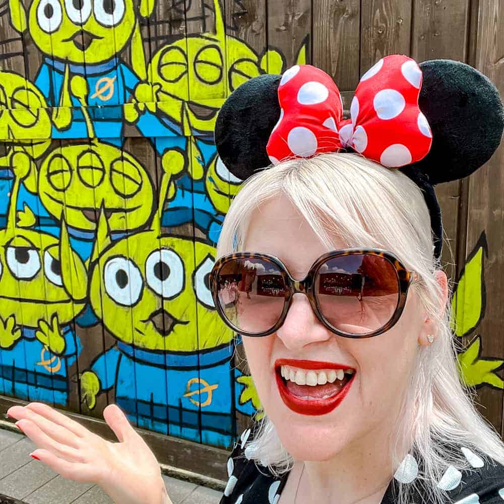 Carrie-Anne, blogger at Crepes and Castles, wearing Minnie ears and sunglasses at Disneyland Paris in June, smiling in front of the Toy Story aliens mural in Walt Disney Studios Park