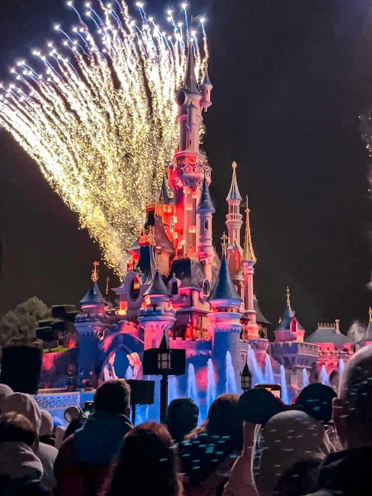 Nighttime fireworks show at Sleeping Beauty Castle during June at Disneyland Paris, with fountains and a crowd enjoying the performance