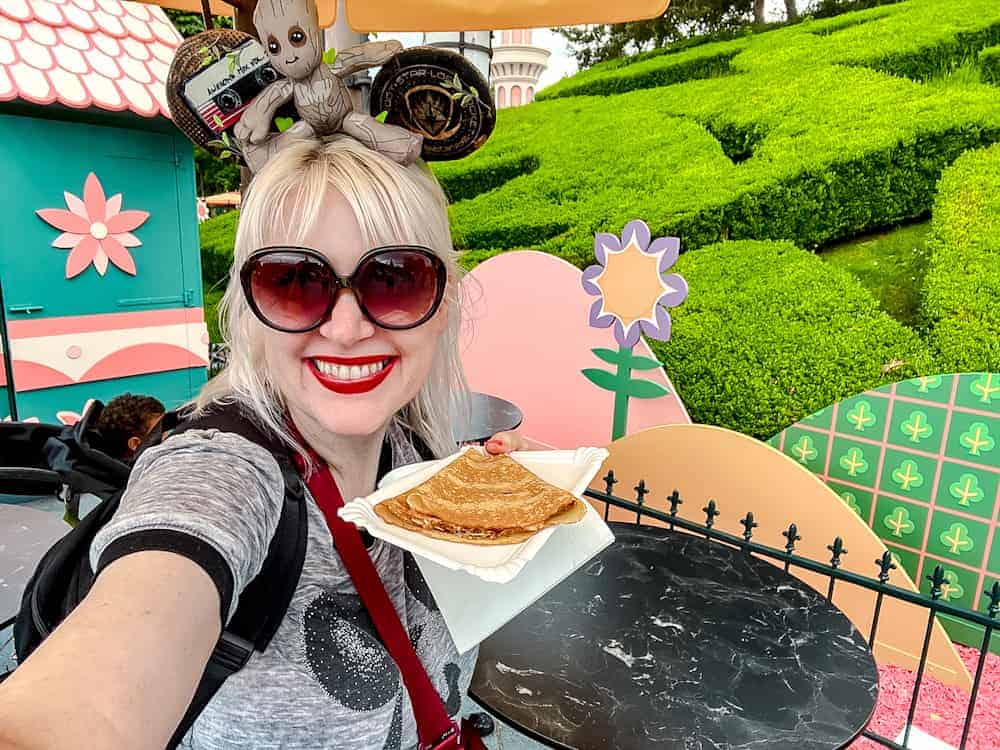 Blogger Carrie-Anne smiles with a crêpe in front of colourful Alice in Wonderland scenery in May at Disneyland Paris