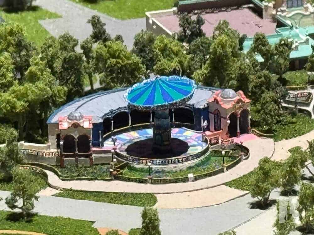 A detailed model of the upcoming Up-inspired swing ride at Disneyland Paris, featuring a colourful canopy, whimsical architecture, and a circular platform surrounded by lush greenery and decorative fencing.