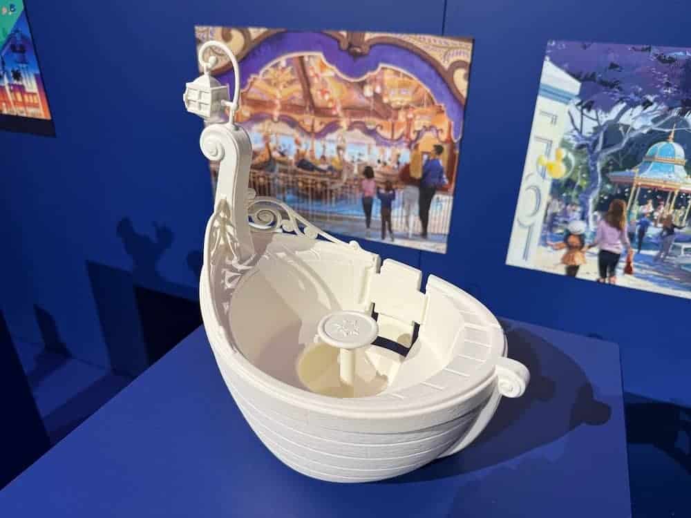 A white model of the Tangled ride boat on display, showing the detailed design including lantern-style lighting and curved seating, with artwork in the background.