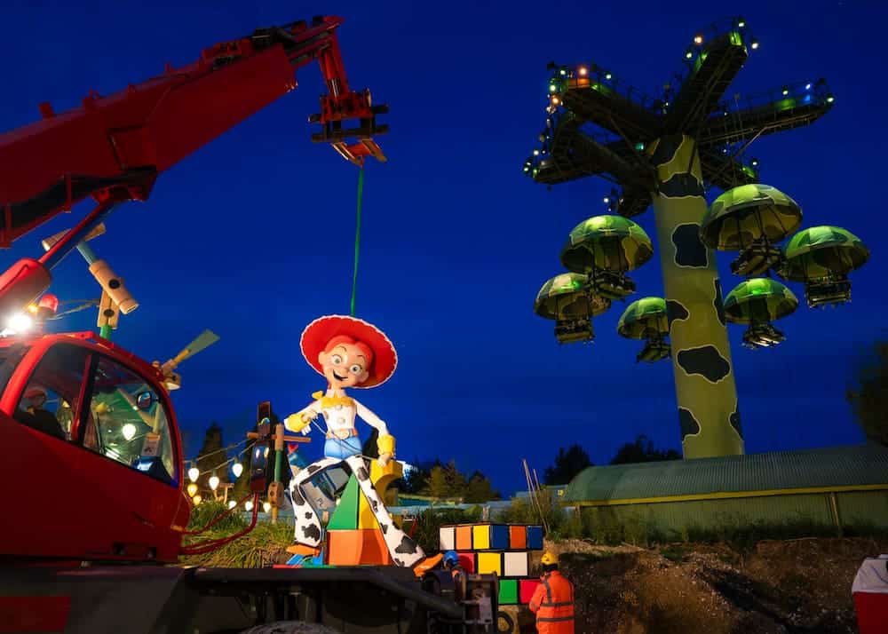 Nighttime photo of a large Jessie figure being installed in Toy Story Playland at Disneyland Paris, with the Toy Soldiers Parachute Drop lit up in the background.
