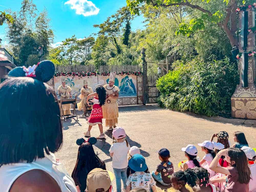 Moana performing with Polynesian drummers at the Disney Music Festival, as children watch from the front rows. A sunny outdoor show full of charm.