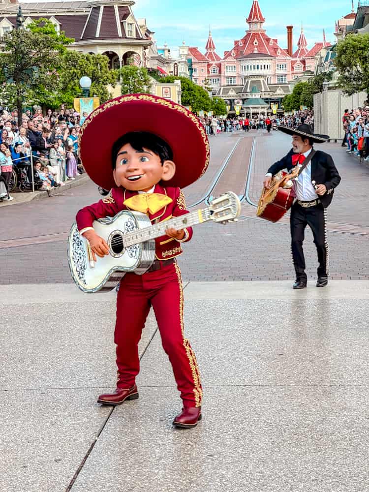 Miguel from Coco performs with his guitar at the Disney Music Festival, with the iconic Disneyland Hotel in the background. A joyful moment on Main Street.