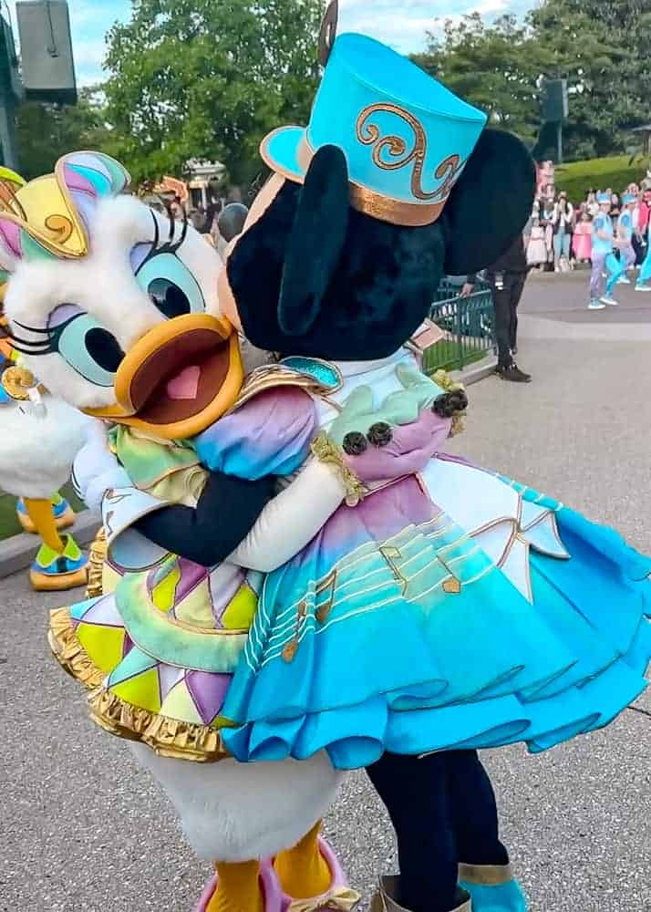 Daisy Duck and Minnie Mouse embrace during the Disney Music Festival at Disneyland Paris, both dressed in colourful, music-themed outfits. A heartwarming moment between friends.