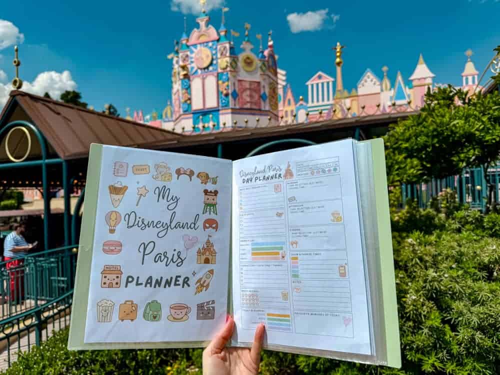 A colourful Disneyland Paris planner from Crepes and Castles is held open in front of ;it’s a small world,' showing a cute daily itinerary page and kawaii-themed cover design. A helpful tool for navigating the Disney Music Festival.
