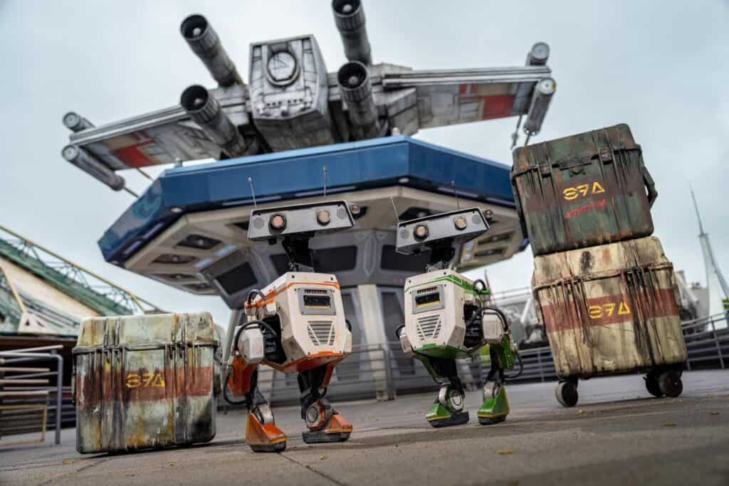 Two BDX droids—one orange, one green—pose playfully in front of crates and the towering X-Wing above Lounge Lightspeed at Disneyland Paris. The droids are boxy, mechanical, and full of personality, with antennas raised like curious ears.