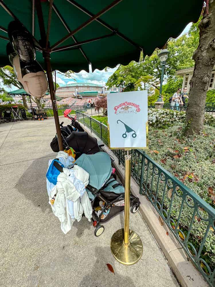 Pushchair parking area near Plaza Gardens restaurant at Disneyland Paris, showing multiple strollers under shaded umbrellas.