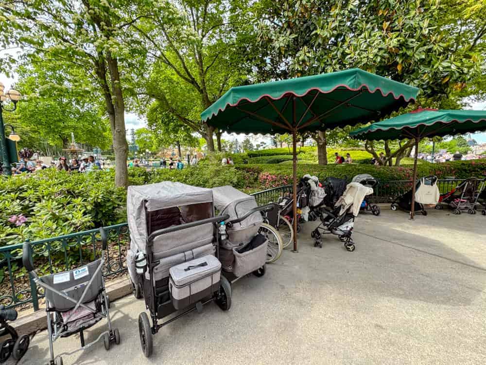 Several large pushchairs parked under green umbrellas in a stroller zone at Disneyland Paris near the hub.