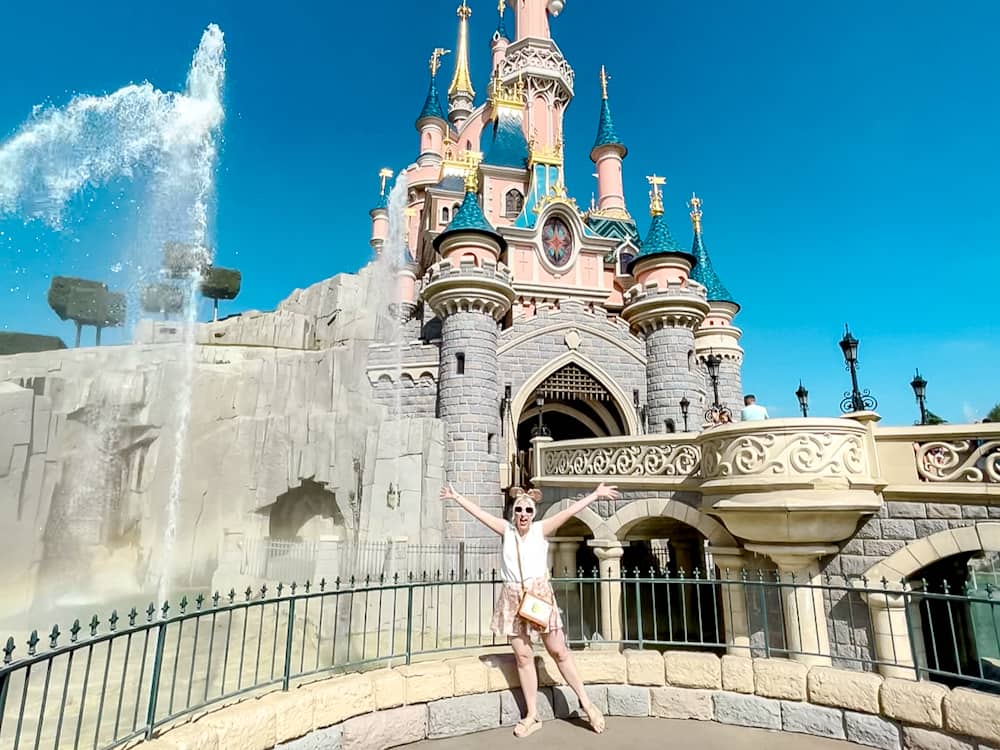 Excited visitor poses in front of Sleeping Beauty Castle as fountains spray — a highlight of August at Disneyland Paris