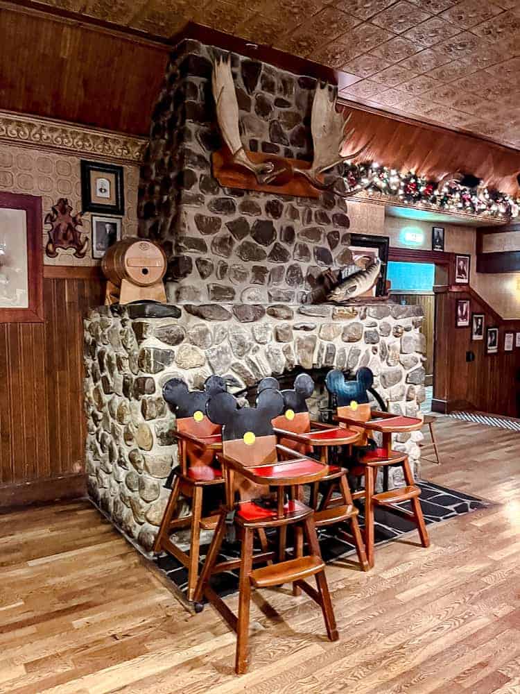 A group of wooden high chairs with Mickey Mouse ears in front of a stone fireplace at Billy Bob’s La Grange, Disneyland Paris.