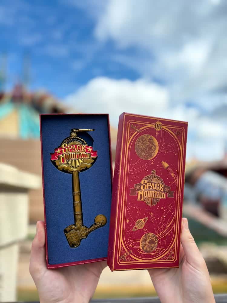 Close-up of the Disneyland Paris Hyperspace Mountain 30th anniversary collectible key in red and gold packaging, with a clear sky and ride structure in the background.