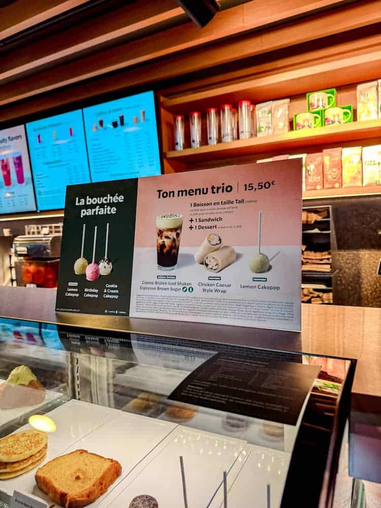 Close-up of the Ton Menu Trio sign at Starbucks in Disney Hotel Cheyenne, showing the €15.50 deal with a drink, sandwich, and dessert, including options like donuts and cake pops.