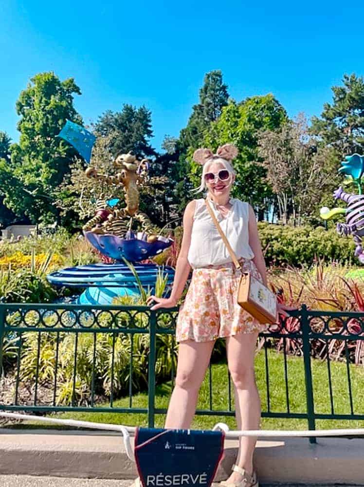 Carrie-Anne in Minnie ears and floral shorts posing by the Castle hub statues during a sunny day in July at Disneyland Paris.