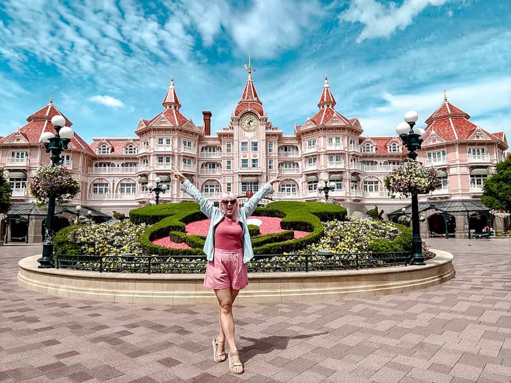DLP blogger Carrie-Anne Hall in pink shorts with Mickey ears and arms raised in front of the Disneyland Paris Hotel – a joyful moment for first-timers making full use of these Disneyland Paris planning tips!