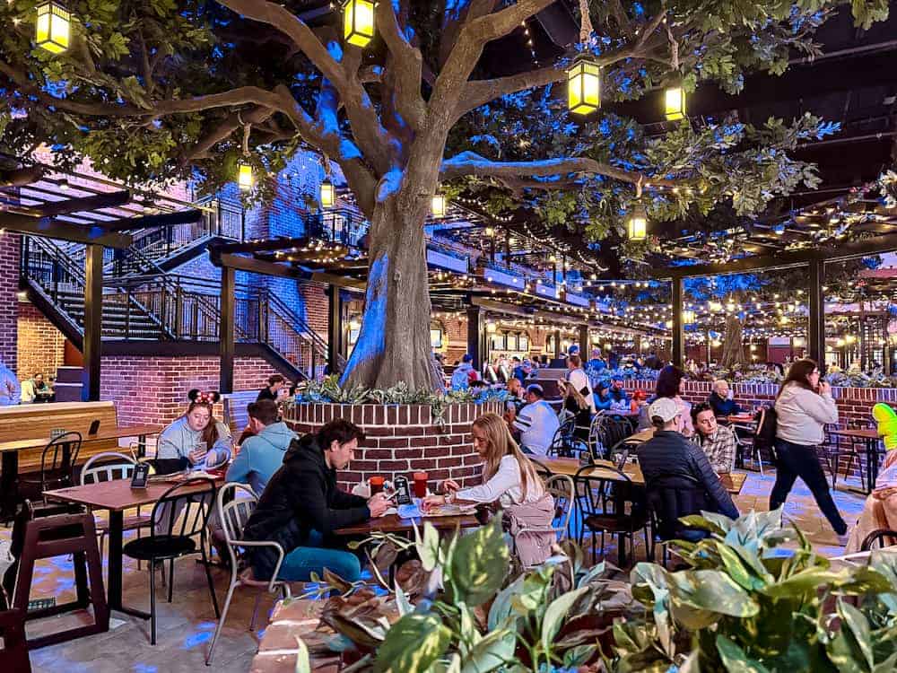 Diners seated beneath a large tree with glowing lanterns inside Hollywood Gardens Restaurant at World Premiere Disneyland Paris.