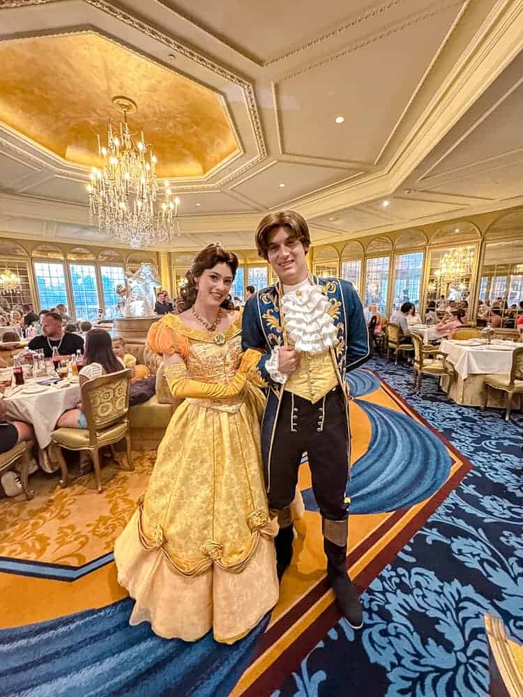 Belle and her prince posing inside La Table de Lumière at Disneyland Paris, with chandeliers and guests dining in the background.