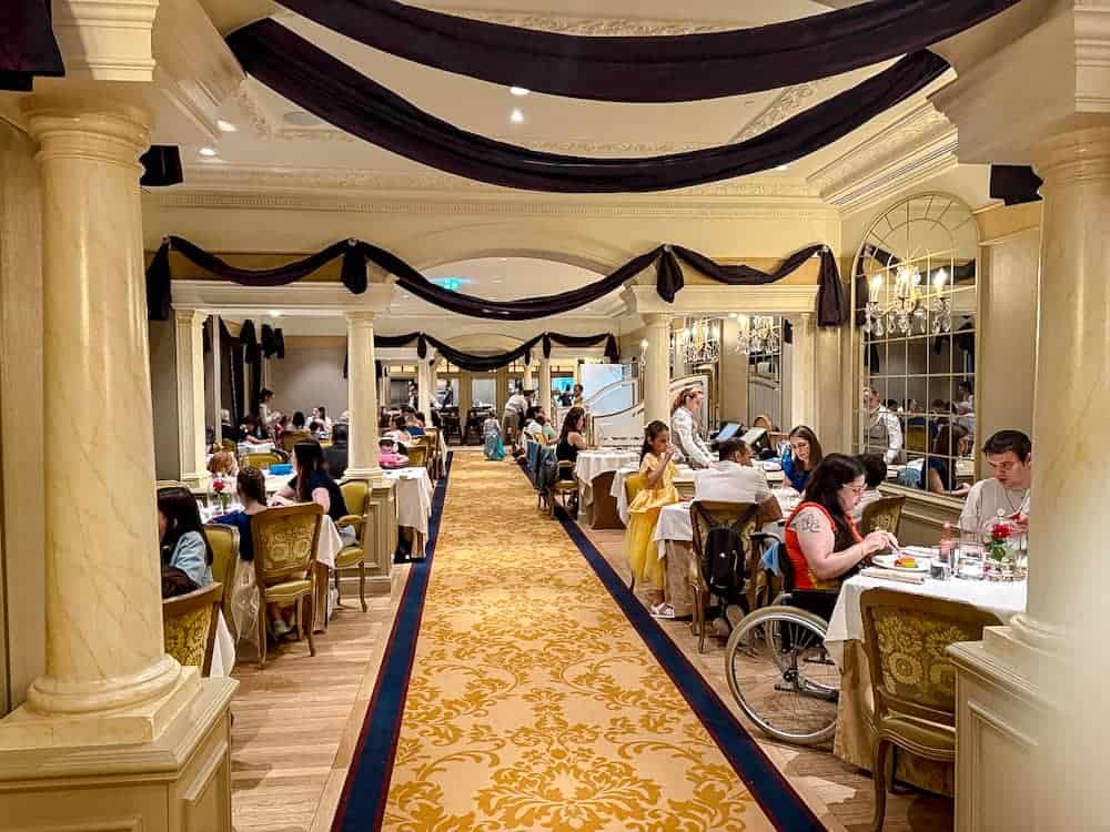 The hallway dining section at La Table de Lumière in Disneyland Paris, with golden chairs, chandeliers, and black draped fabric adorning the ceiling.