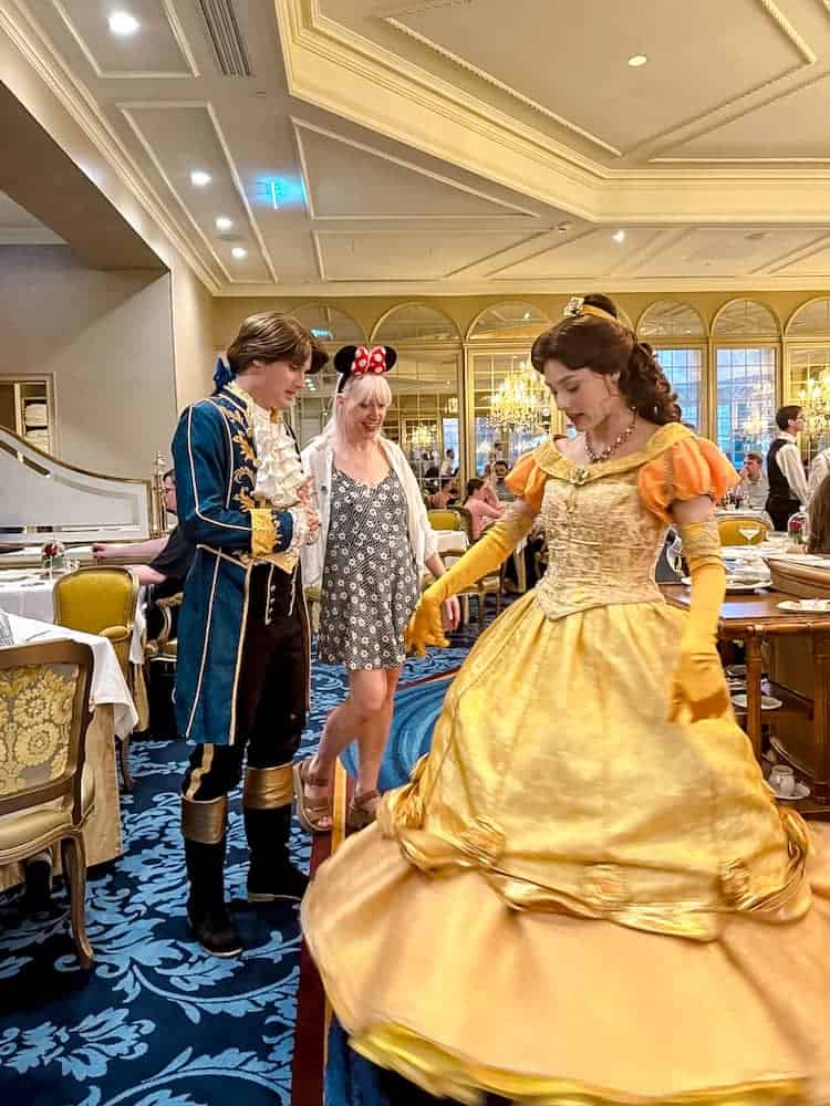 Princess Belle swishes her golden gown mid-twirl while Prince Adam chats with a guest wearing Minnie ears at La Table de Lumière in Disneyland Paris.
