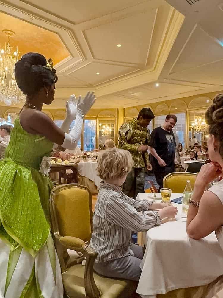 A young child at a dining table being greeted by Princess Tiana in her green gown, with Prince Naveen in the background, at La Table de Lumière inside the Disneyland Hotel.