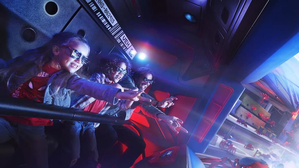 A family wearing 3D glasses on Spider-Man W.E.B. Adventure at Disneyland Paris, reaching out to sling webs at digital Spider-Bots during the interactive ride experience