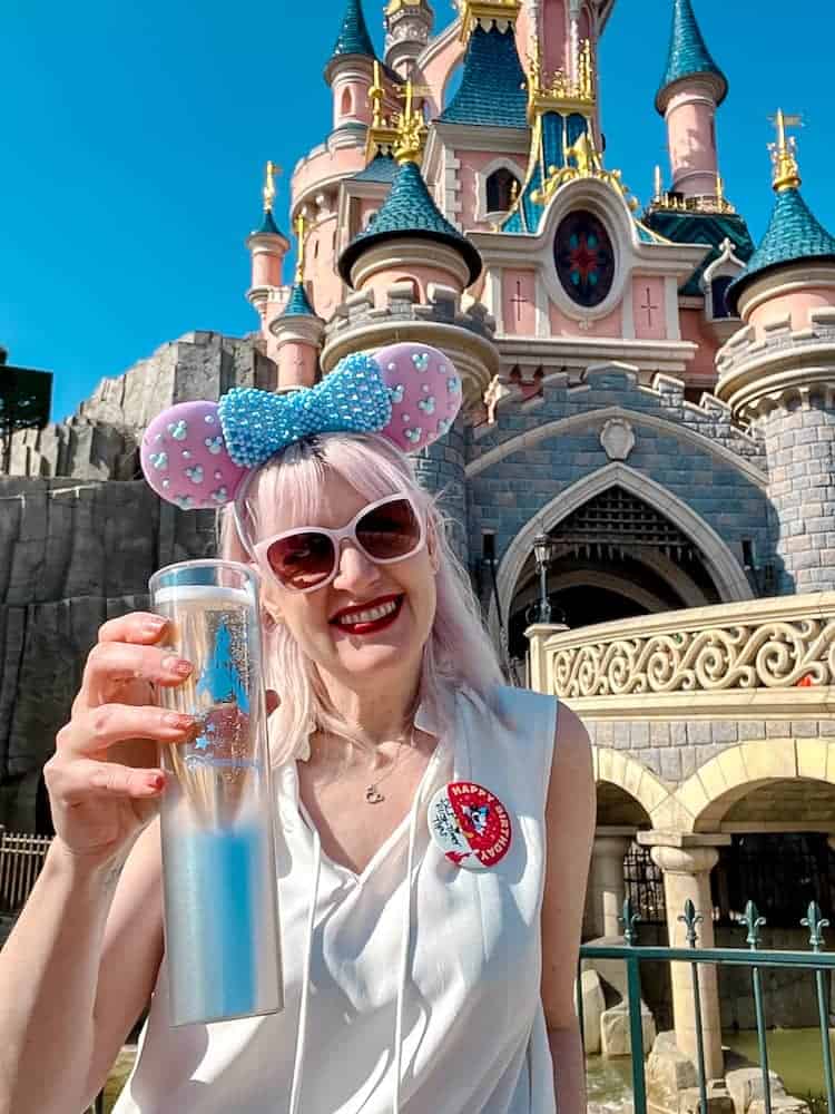 Smiling visitor with Minnie ears holding champagne in front of Sleeping Beauty Castle, celebrating arrival at Disneyland Paris after French Air Traffic Control Strike September 2025.