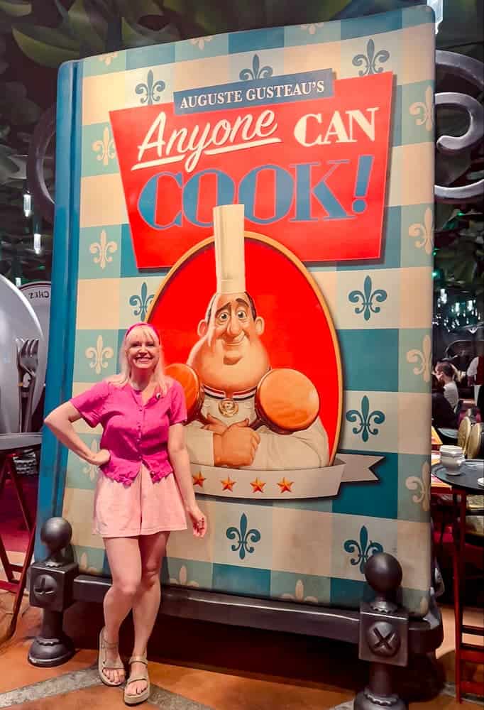 Disney blogger Carrie-Anne posing in front of giant “Anyone Can Cook!” cookbook inside Bistrot Chez Rémy at Disneyland Paris