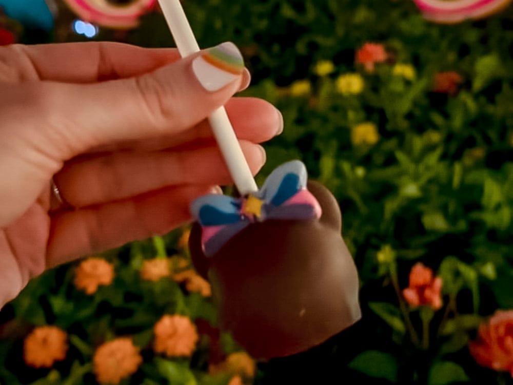 A manicured hand holds a chocolate covered Disneyland California cake pop themed for the 70th anniversary with colourful Minnie bow