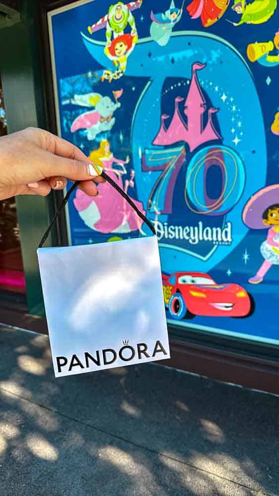 Hand holding a Pandora shopping bag in front of a colourful Disneyland 70th Celebration sign with Disney characters in the background.