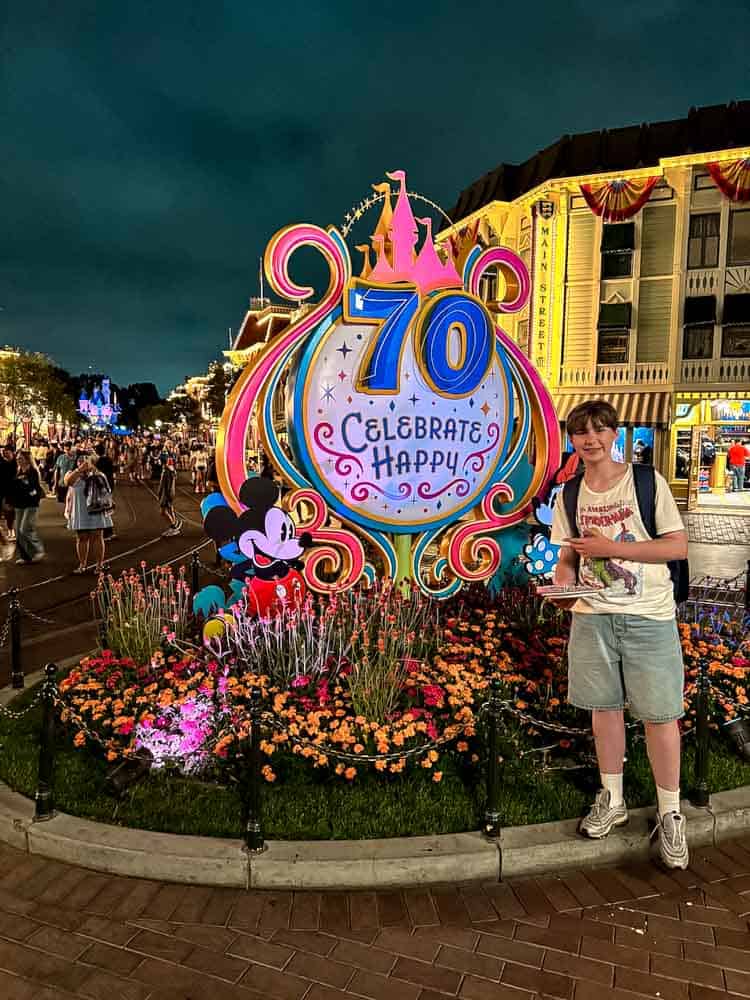 Teen boy posing with a churro by the Disneyland 70th Celebration sign on Main Street at night during a 3-Day Disneyland California Itinerary.