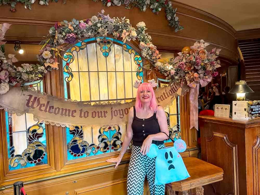 Disneyland Paris blogger Carrie-Anne Hall standing at the Unlucky Nugget entrance surrounded by floral garlands and a 'Welcome to our joyous celebration' banner at Disneyland Paris Halloween 2025.