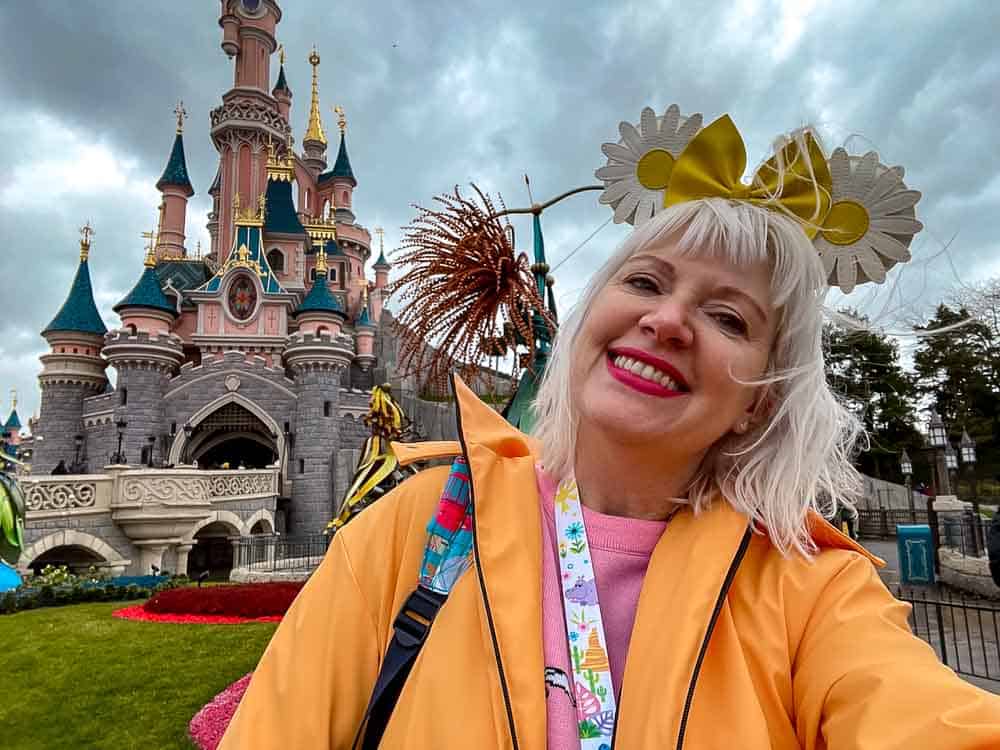 Carrie wearing a bright orange waterproof coat and daisy Minnie ears in front of Sleeping Beauty Castle on a stormy Disneyland Paris day.