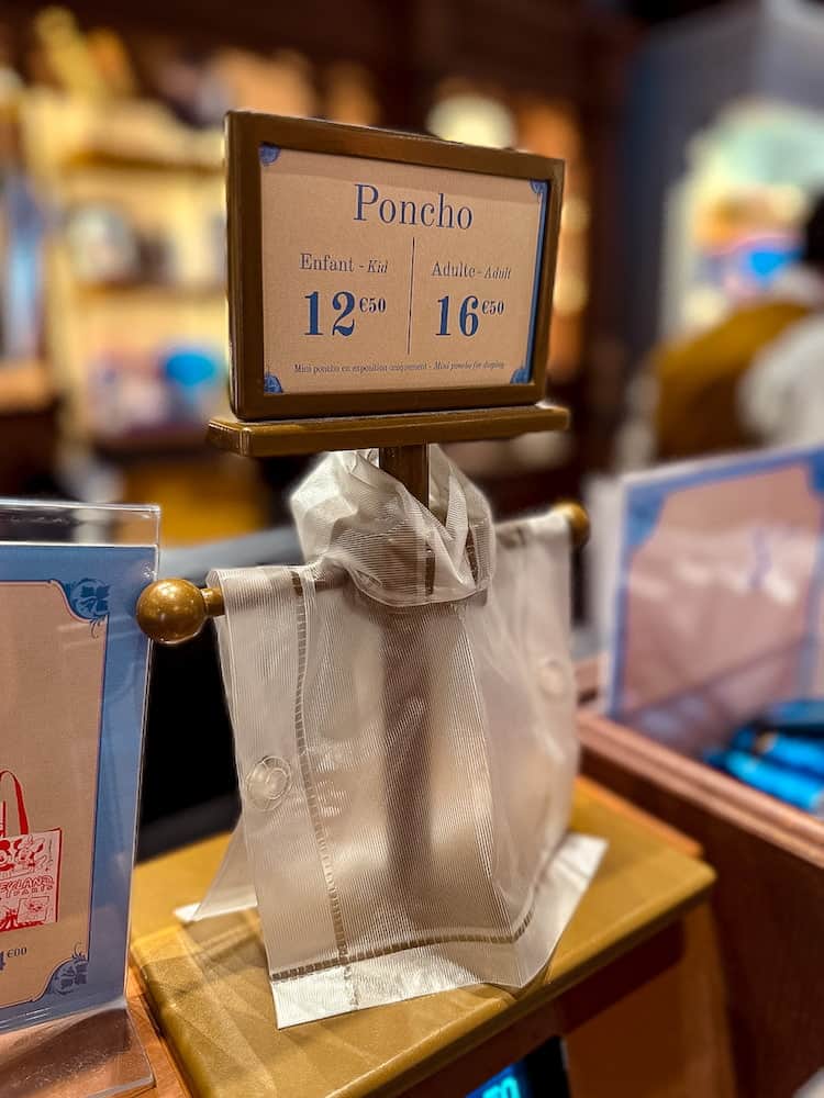 Close-up of Disneyland Paris poncho prices showing €12.50 for kids and €16.50 for adults, with a mini display poncho at the till.
