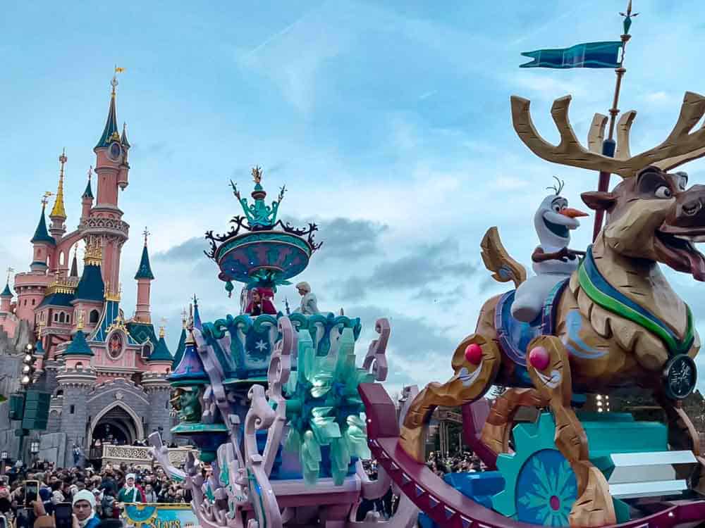 Olaf and Sven on a Frozen-themed parade float with Sleeping Beauty Castle in the background, under a cloudy sky at Disneyland Paris.