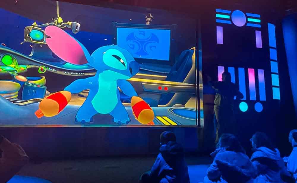Children watching the Stitch Live! show inside Walt Disney Studios Park at Disneyland Paris in the rain, with Stitch on screen interacting with the audience