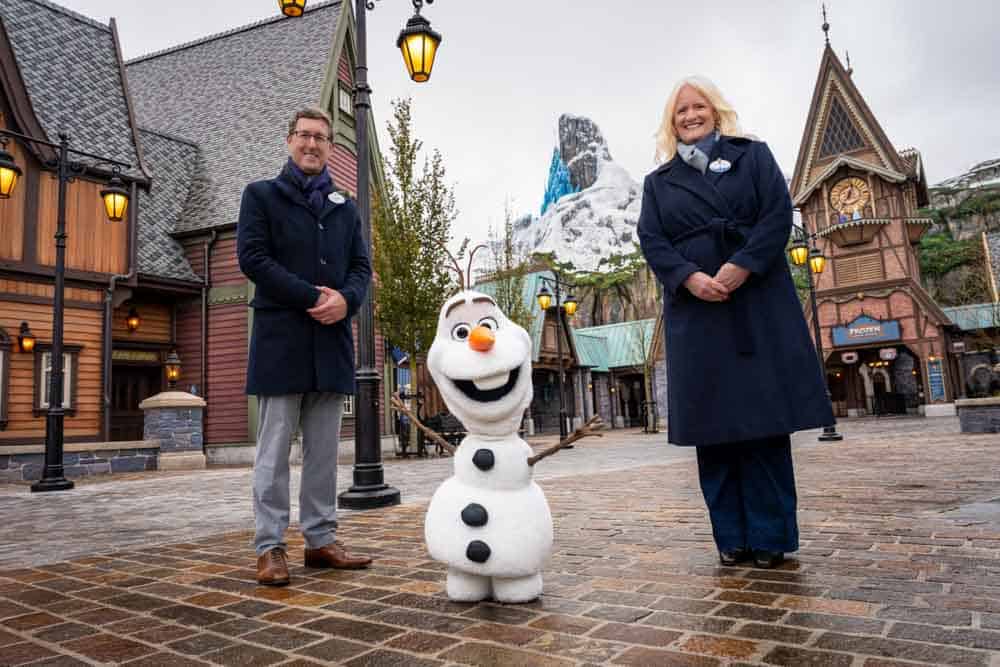Olaf standing with Disney Imagineering leaders in the village area of World of Frozen at Disneyland Paris with the North Mountain in the background.