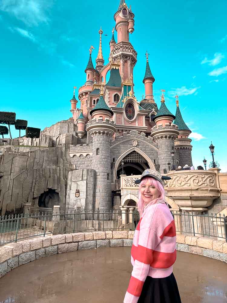 Disneyland Paris blogger Carrie-Anne Hall smiling in front of Sleeping Beauty Castle, wearing a pink-and-white outfit on a bright blue-sky day.