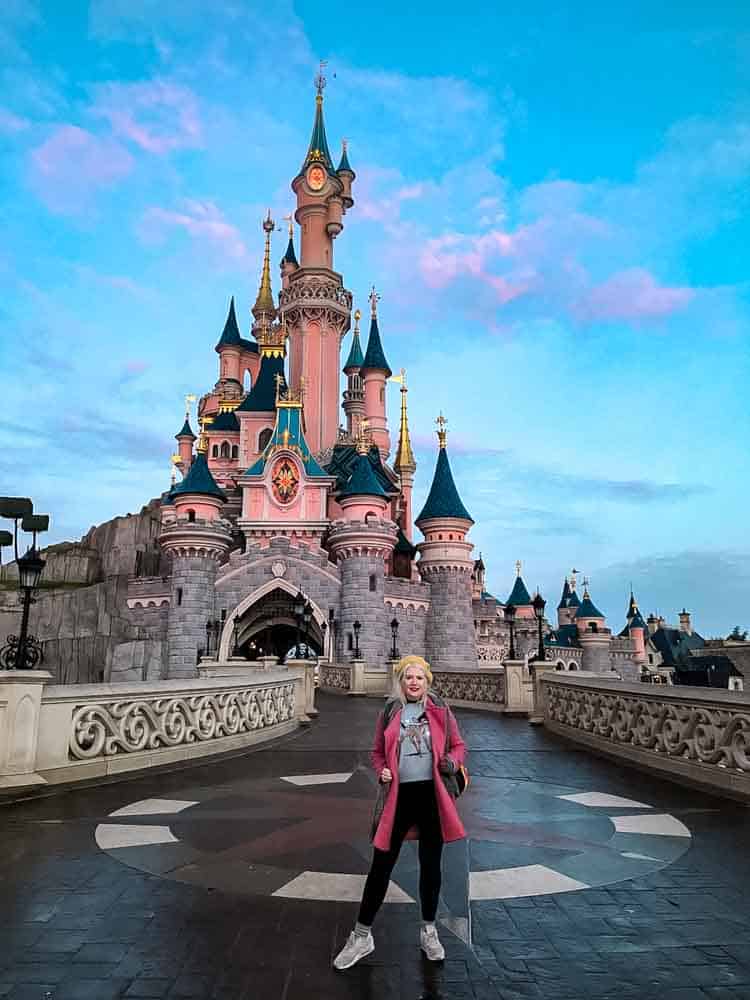 Sleeping Beauty Castle at Disneyland Paris at sunset in January, with visitor posing in the foreground.
