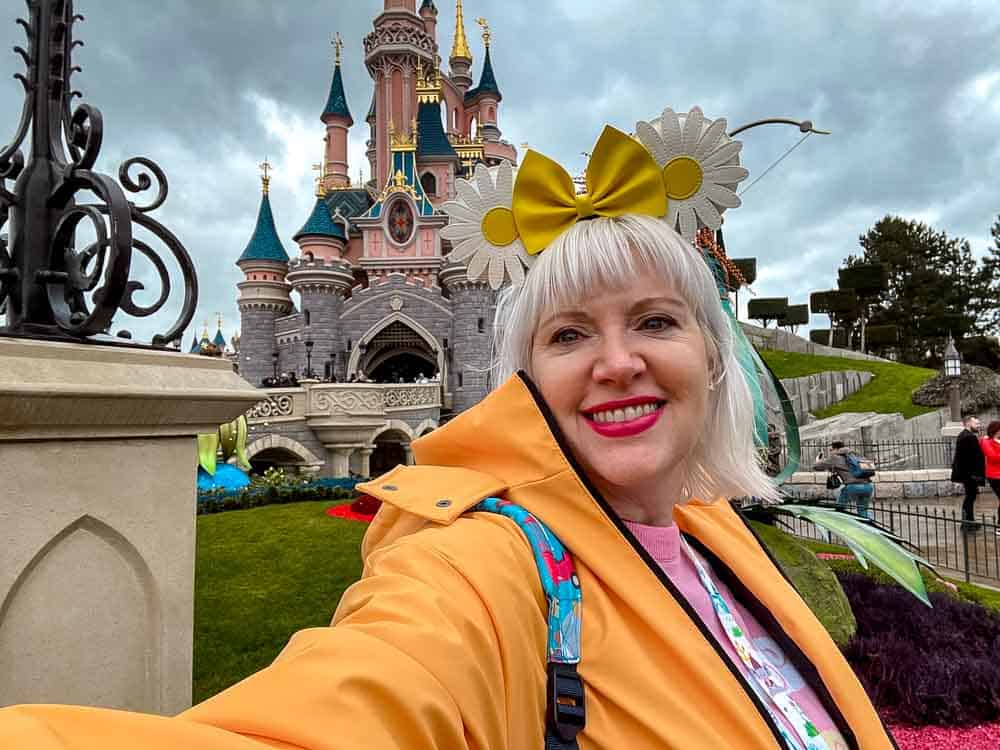 Disney blogger Carrie-Anne visiting Disneyland Paris in March wearing a raincoat, with Sleeping Beauty Castle in the background on a cool, cloudy day