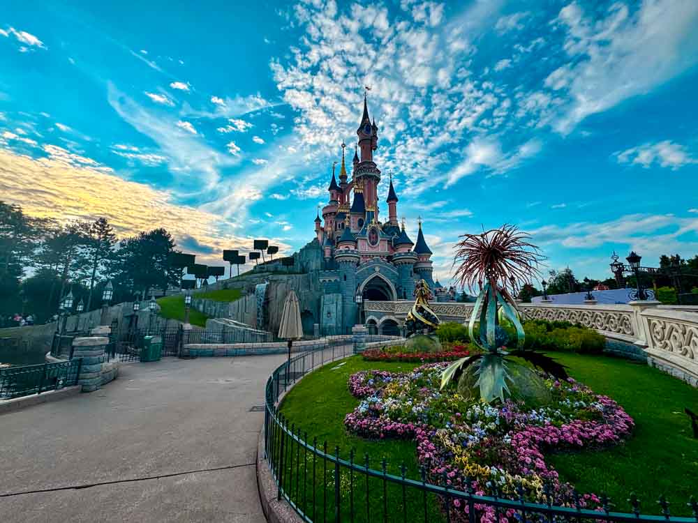 Best Disneyland Paris Castle Photo Spots include this wide view of Sleeping Beauty Castle from the left side path at sunrise, with circular flower bed and ornamental sculpture in foreground