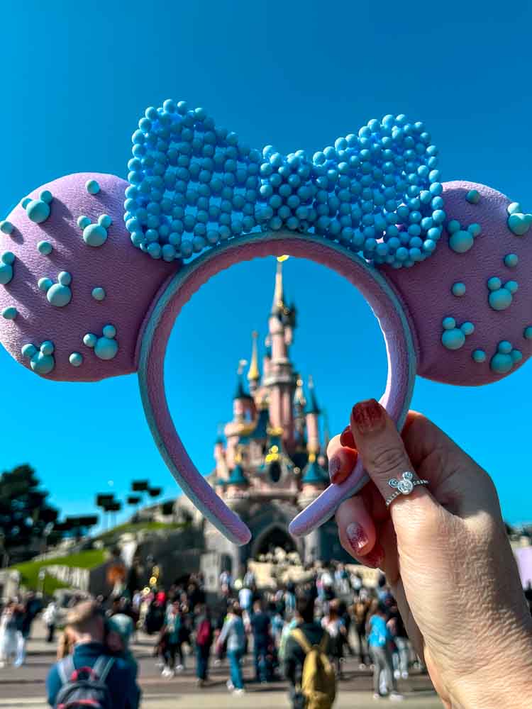Blue and pink Minnie Mouse ears held up to frame Sleeping Beauty Castle against a bright blue sky at Disneyland Paris