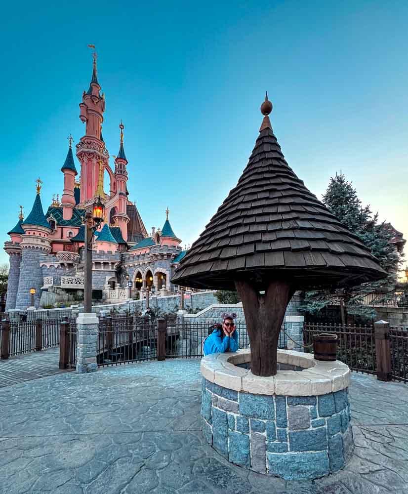 Woman leaning on the Snow White wishing well on the right side of Sleeping Beauty Castle at Disneyland Paris, castle visible behind in early morning light