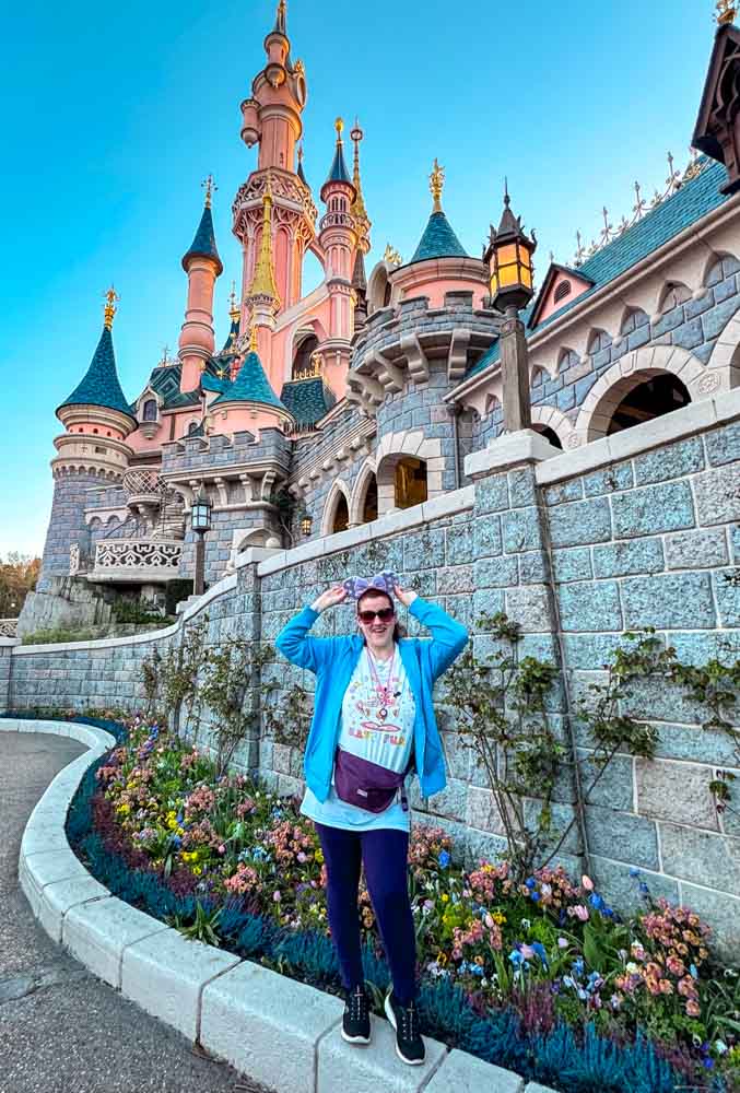Woman standing in front of the curved flower beds along the right side castle wall at Disneyland Paris, castle towering behind her