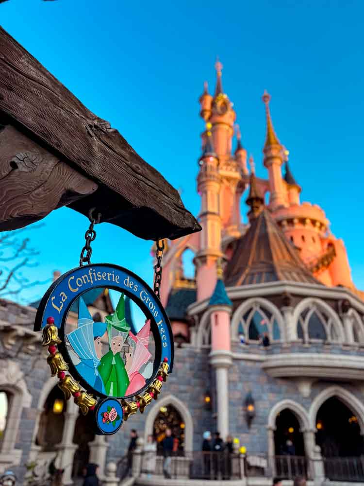 La Confiserie des Trois Fées decorative shop sign in Fantasyland at Disneyland Paris, with Sleeping Beauty Castle glowing at dusk behind it